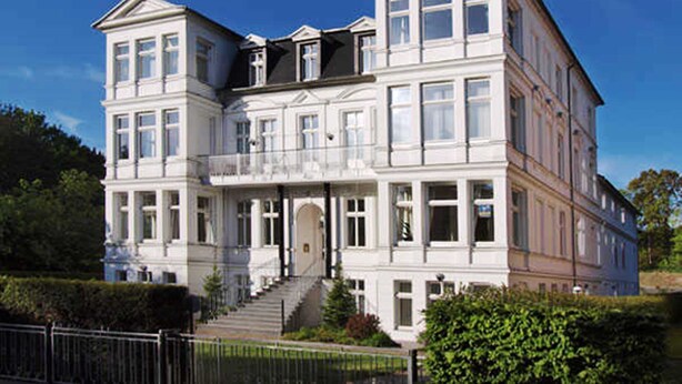 Un grand bâtiment blanc à plusieurs étages, doté d’une façade symétrique, de nombreuses fenêtres et d’une entrée centrale avec des marches menant à celle-ci.