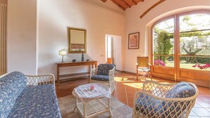 Habitació acollidora amb sostre de fusta, un finestral gran amb vistes a vegetació, un sofà de vimet, una taula petita amb una làmpada, i una cadira amb coixí.