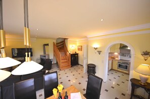 A dining area with a wooden table, chairs, and pendant lights. A staircase leads to a kitchen area with a stove and cabinets.