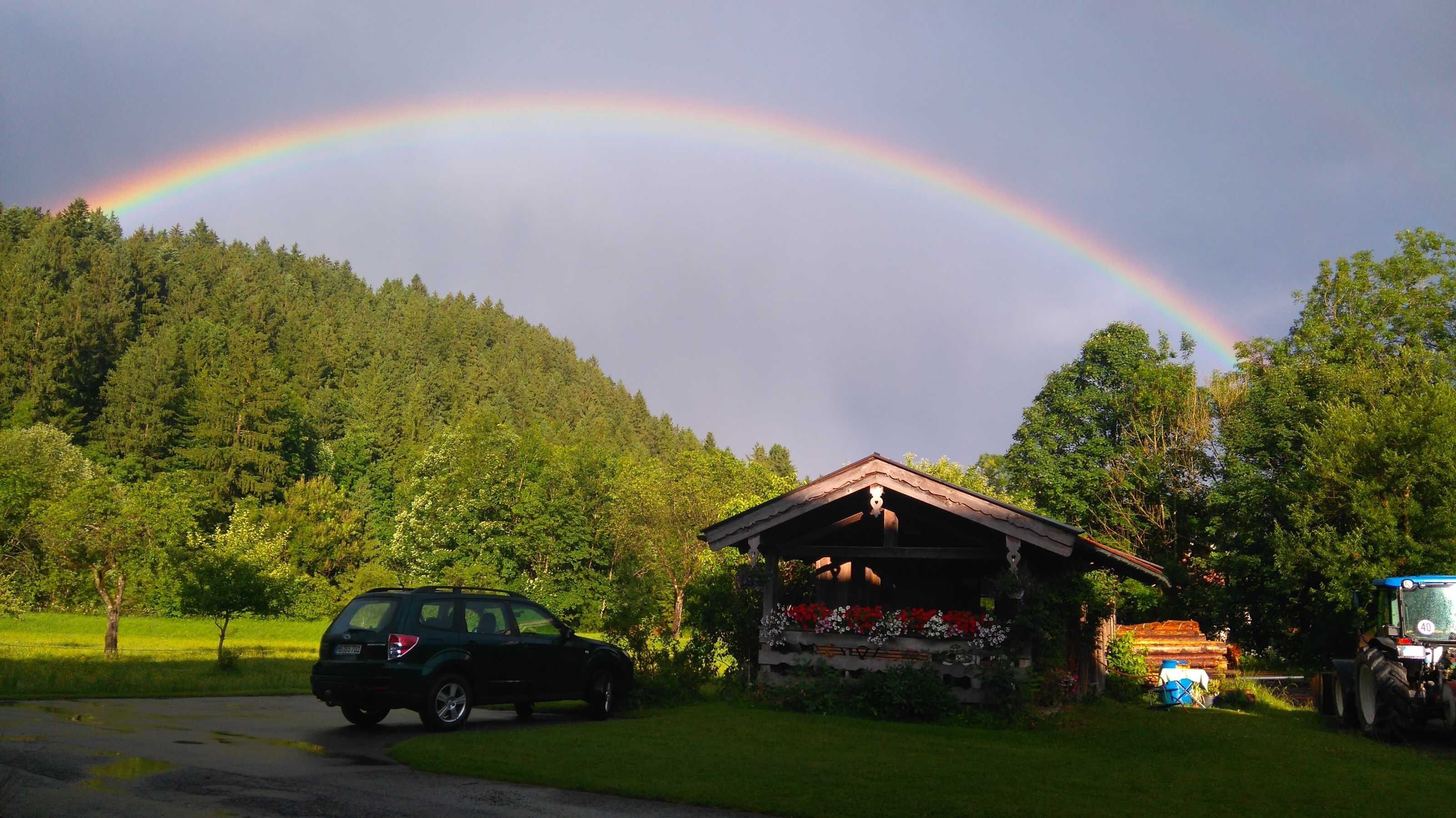 Ländliche Szenerie mit Regenbogen, einem kleinen Holzhaus, einem geparkten Auto und einem Traktor.