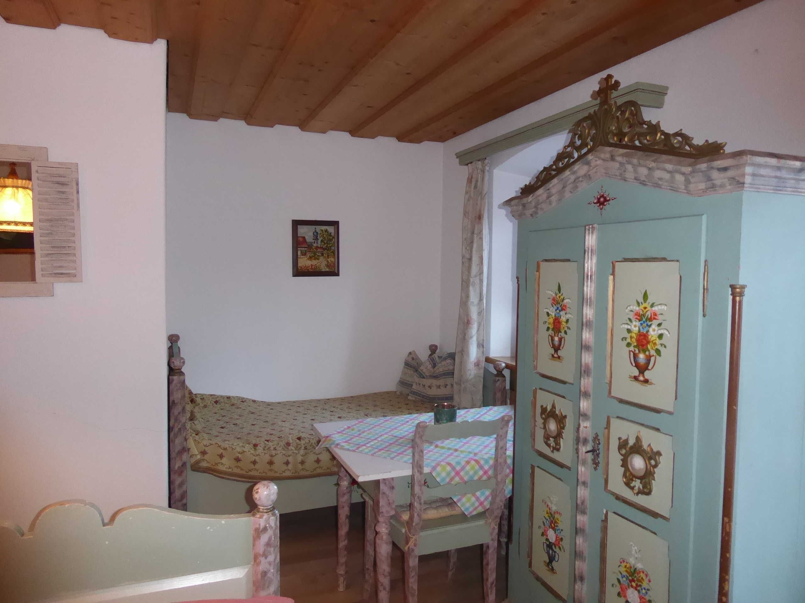 Ein Zimmer mit einem Bett, einem Tisch mit kariertem Tischtuch, einem Schrank mit Verziertungen und einer Holzboden.