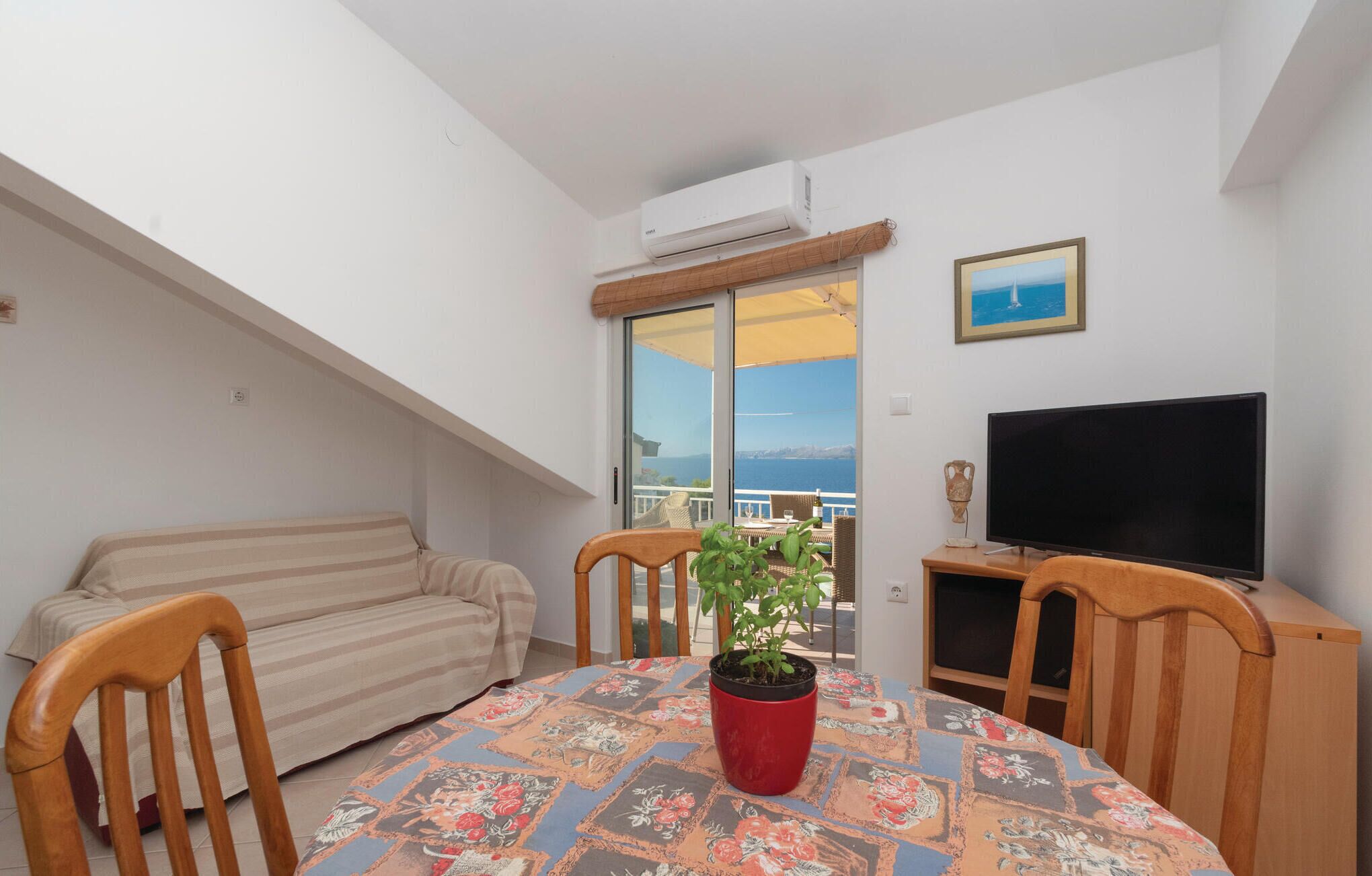 A balcony with a sofa, a dining table with chairs, a potted plant, a flat-screen TV, and a framed picture on the wall.