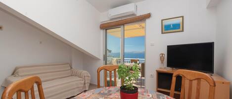A balcony with a sofa, a dining table with chairs, a potted plant, a flat-screen TV, and a framed picture on the wall.