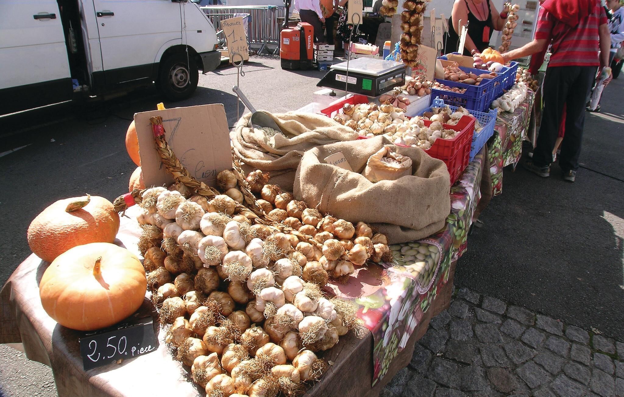 Un puesto de mercado con diversas verduras como cebollas y calabazas, una balanza y un cartel con los precios.