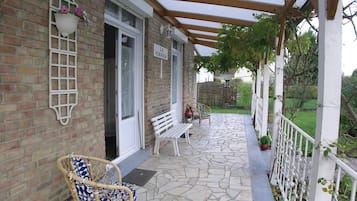 Een overdekt terras met een bank en een rieten stoel, een bakstenen muur en een bordje met de tekst 'LA PERGOLA'.