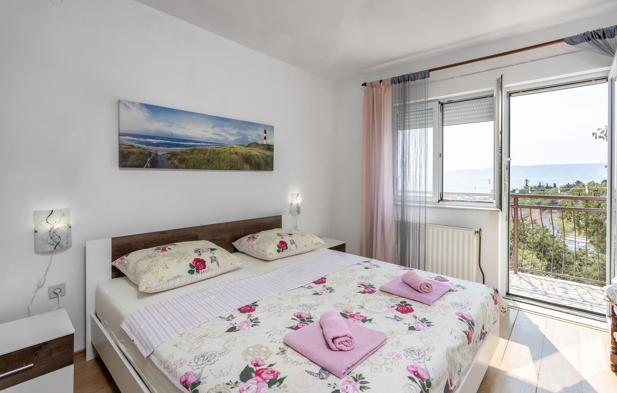 A bedroom with a bed, floral bedding, a painting of a beach, a window with curtains, and a view of greenery.