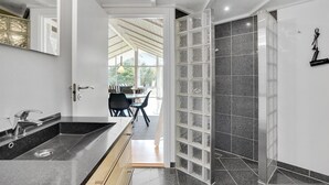 Un baño moderno con un lavamanos grande, cabina de ducha con mampara de vidrio y vista a un área de comedor al aire libre.