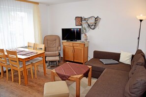 A living room with a wooden dining table, chairs, a sofa, a television, and a lamp. - Ferienappartement 01 - Pension in Ostseebad Sellin (Sellin)