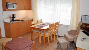 Una cocina compacta con armarios de madera, un microondas y una cafetera. Hay una mesa de comedor con sillas y un sofá con televisor.