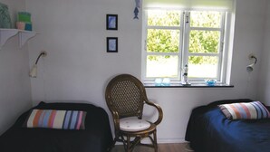 Ein Zimmer mit zwei Betten, einem Sessel, einem Fenster mit Blick auf Grünflächen und einem Regal mit Gegenständen.