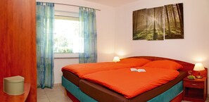 Ein Schlafzimmer mit einem Bett, zwei Nachttischlampen, einem Fenster mit Vorhängen, einem Nachttisch mit Lampe und einer Schale Trauben sowie einer Wand mit einem Waldgemälde.
