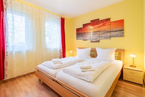 A double bed with white linens, a nightstand with a lamp, and a painting of a sunset on the wall.