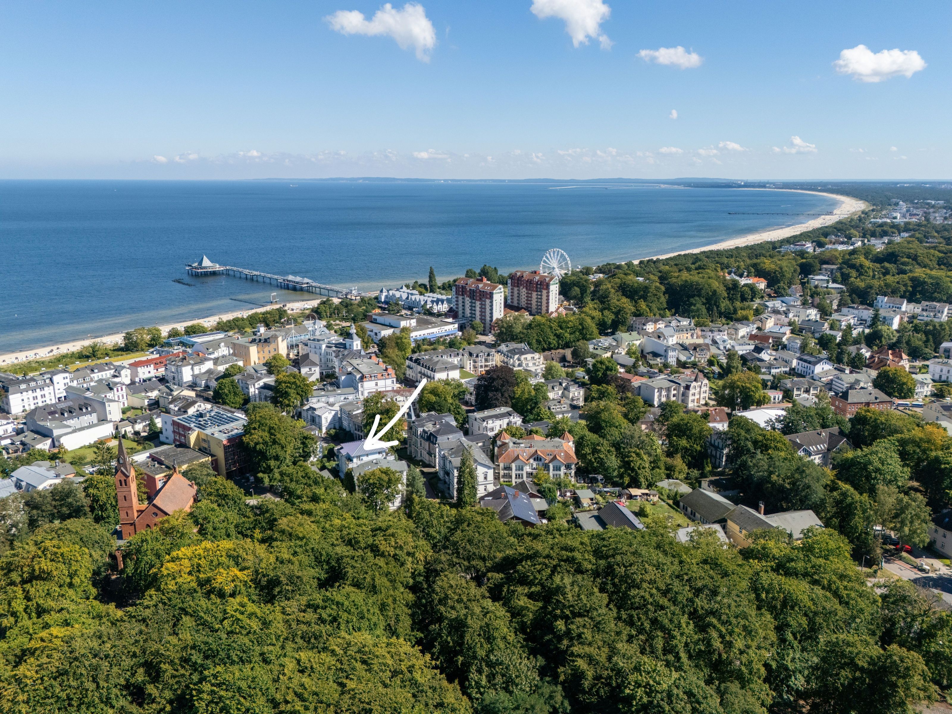 An aerial view of a coastal town with a mix of residential and commercial buildings, a prominent Ferris wheel, and a pier extending into the sea.