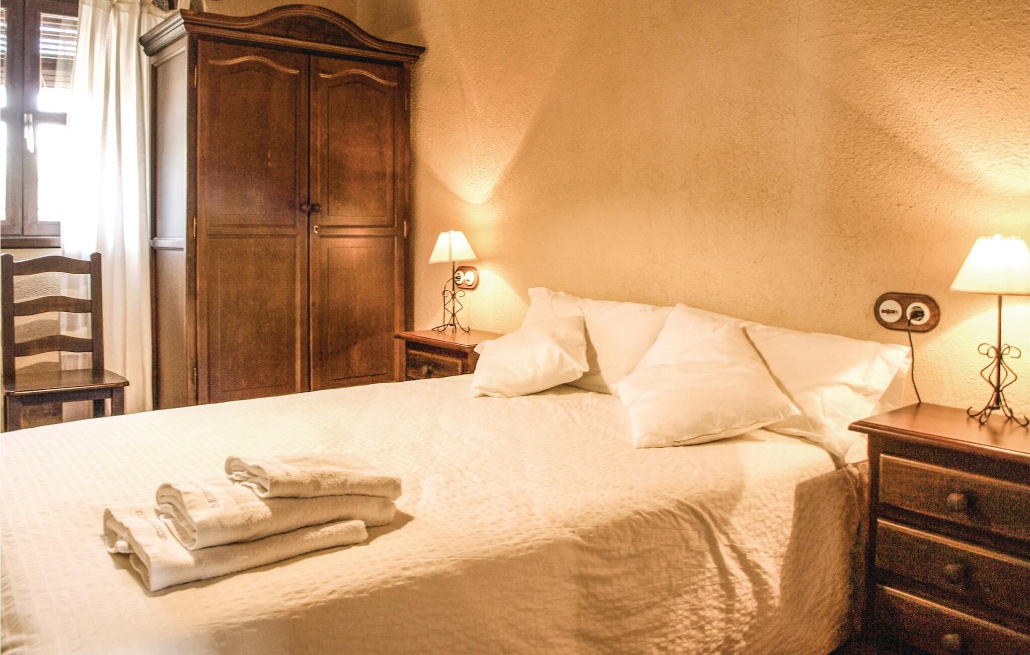 A neatly made bed with white linens and a folded towel, a wooden wardrobe, and two bedside lamps.