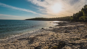 A rocky beach with clear water and a distant tree-covered hill under a bright sky. - Holiday apartment with terrace (Mali Lošinj)