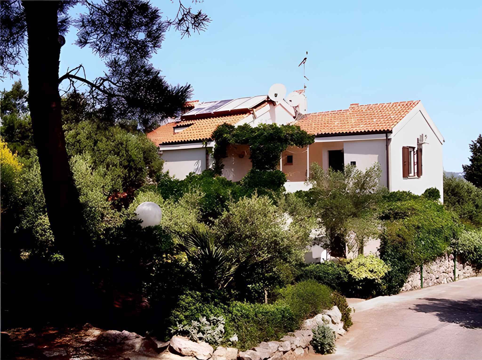 A two-story house with a tiled roof, surrounded by dense greenery and a stone wall.