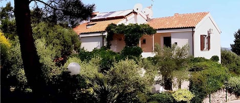 A two-story house with a tiled roof, surrounded by dense greenery and a stone wall.