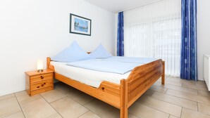 A wooden bed with a nightstand, a framed picture on the wall, and a window with curtains. - Apartment 4 - Landhaus Spittdiek (Neuharlingersiel)