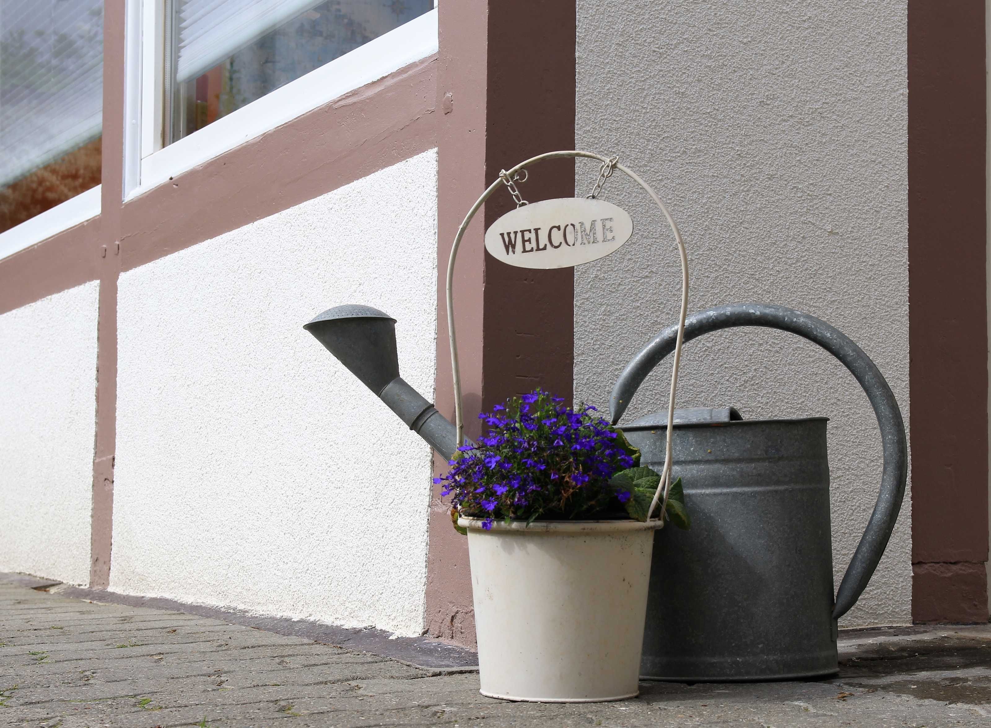 Velkomstskilt, vandkande og blomsterpotte op ad en mur.