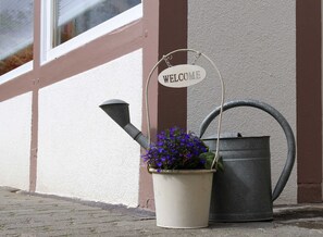 En välkomnande skylt, en vattenkanna och en blomkruka placerade mot en byggnadsvägg.