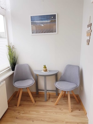 A small room with two grey chairs, a round table, a framed picture of a beach scene, a potted plant, and a sign with room names. - Vacation apartment - Bungalow & vacation apartment Fuchs (Heringsdorf)