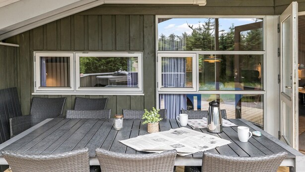 A covered patio with a table set for two, wicker chairs, and a view of the outdoors through large windows. - Gorgeous home in Fjerritslev with sauna (Fjerritslev)