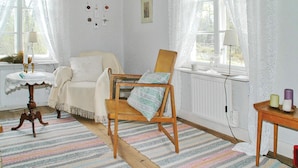 Una habitación con un sillón blanco, una mecedora de madera, un tapete a rayas, una mesita con una lámpara y una ventana con cortinas de gasa.