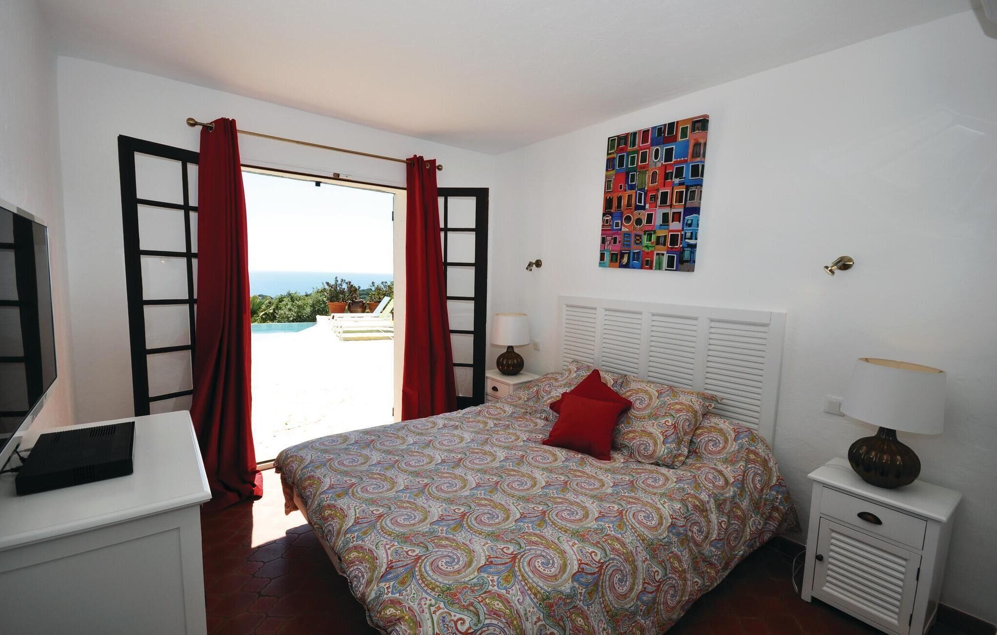 A bedroom with a bed, a desk, a lamp, and a view of the outdoors.