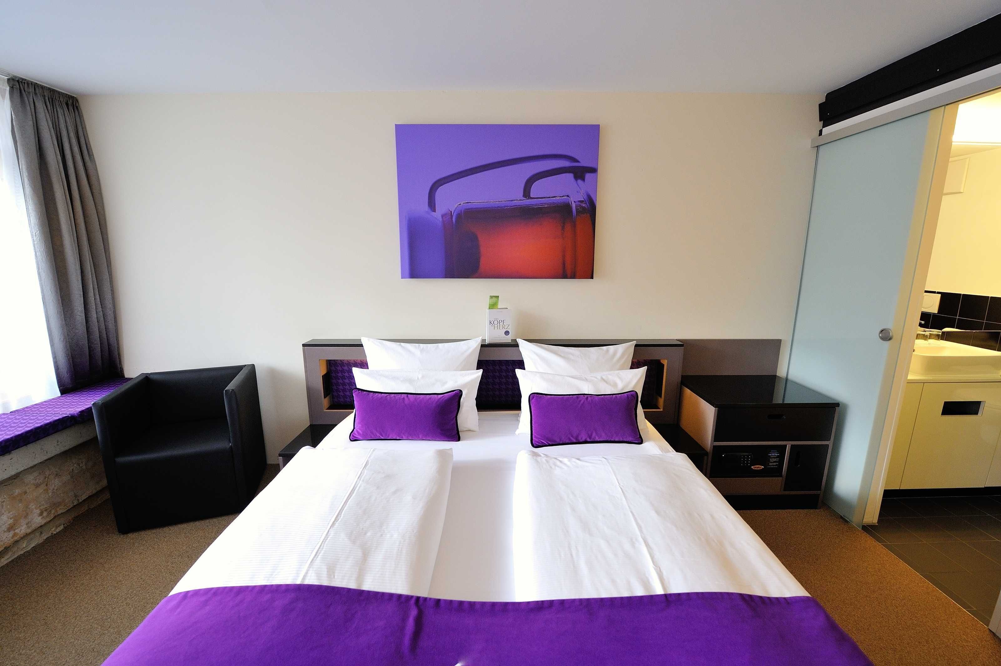Una camera d'albergo con un letto grande, due cuscini, un comodino, una sedia, un quadro appeso al muro e un bagno visibile attraverso una porta aperta.