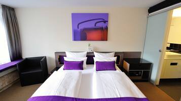 Una camera d'albergo con un letto grande, due cuscini, un comodino, una sedia, un quadro appeso al muro e un bagno visibile attraverso una porta aperta.
