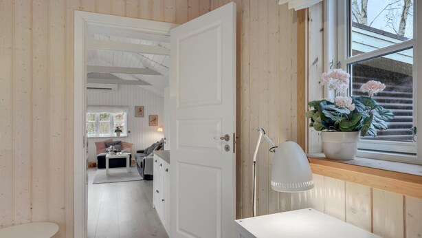 A room with a wooden interior, a white door, a desk, a chair, a window with a view of trees, and a potted plant with flowers. - Amazing home in Nexø with WiFi (Nexø)