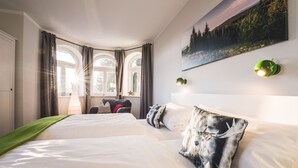 Hotelkamer met een groot bed, twee ramen met gordijnen, een schilderij aan de muur en een groene wandlamp.