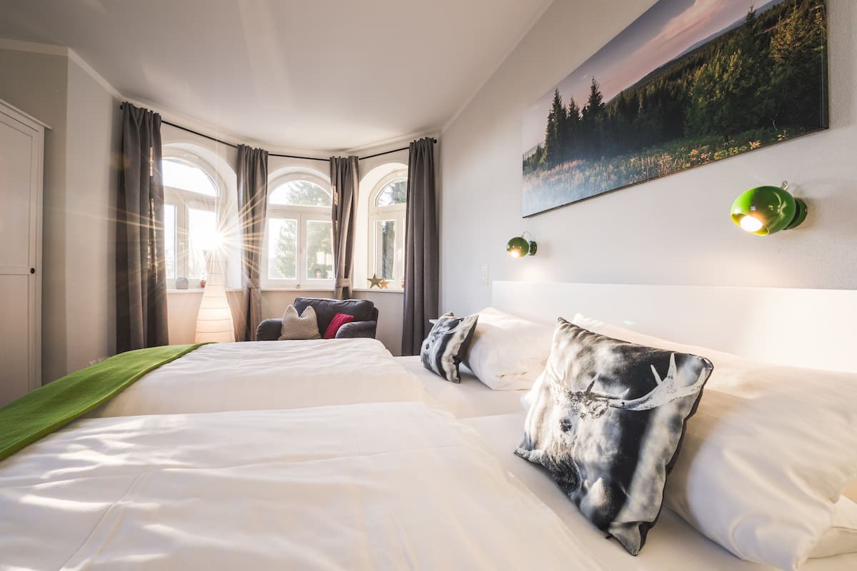 A hotel room with a large bed, two windows with curtains, a painting on the wall, and a green wall lamp.