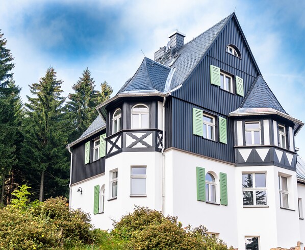 Uma casa grande e escura com detalhes brancos e persianas verdes, rodeada por vegetação exuberante.