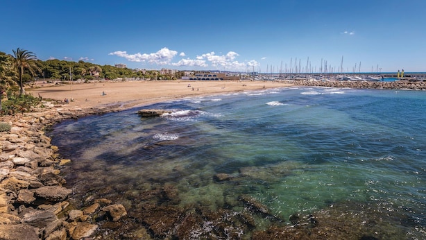 A coastal beach with clear water, rocky shoreline, and a marina with sailboats.