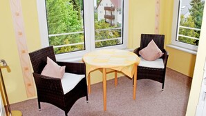 Een hoekzaal met twee rieten stoelen, een klein rond tafeltje en grote ramen met uitzicht op een woonwijk.