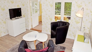 Een kamer met een televisie op een wit kastje, een zwarte stoel, een klein rond tafeltje en een lamp.