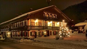 Un edificio de dos pisos con balcón, suelo cubierto de nieve y un árbol de Navidad con luces.