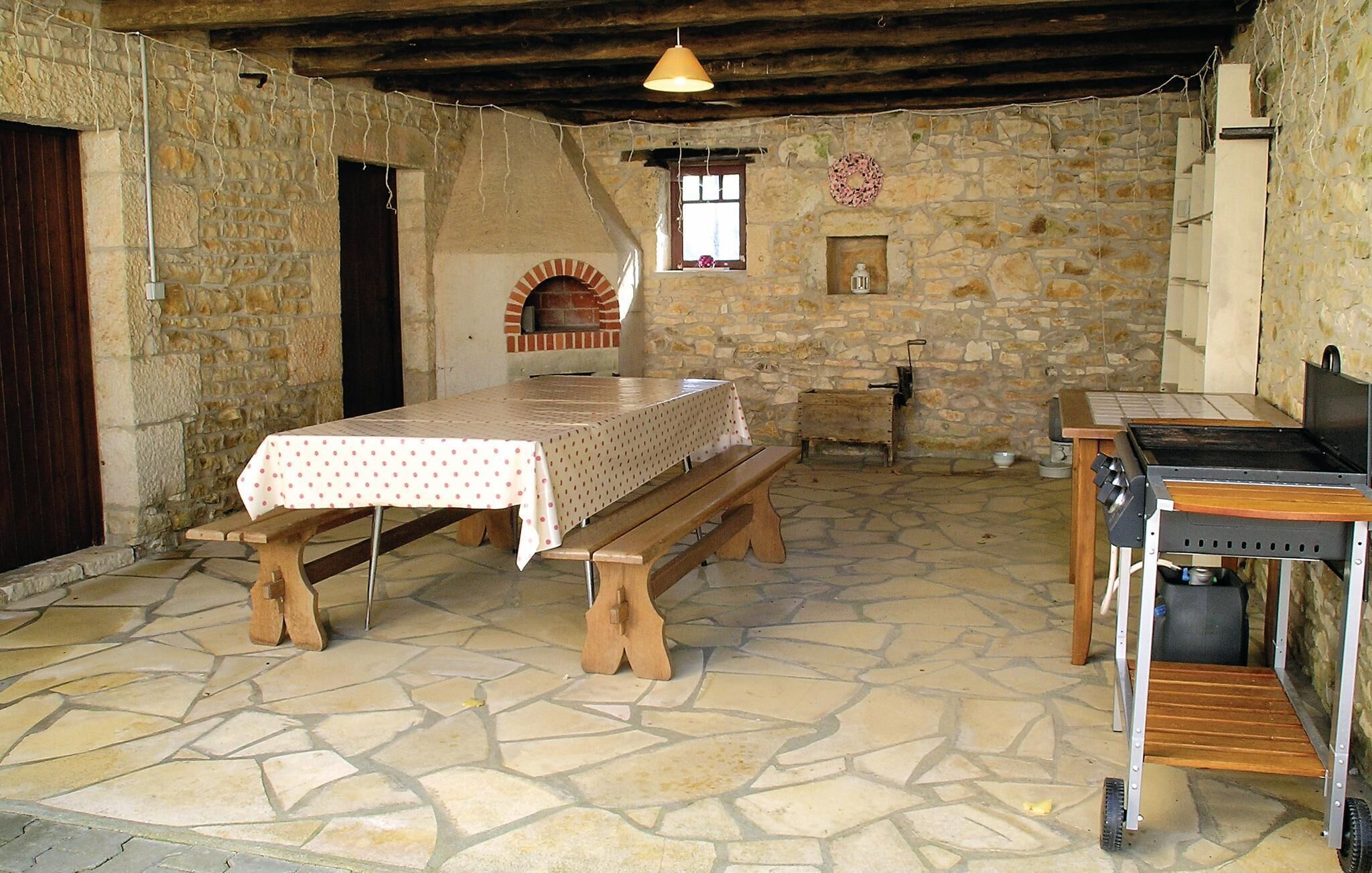 A rustic stone-walled room with a wooden table and benches, a brick oven, and a small window with a flower wreath.