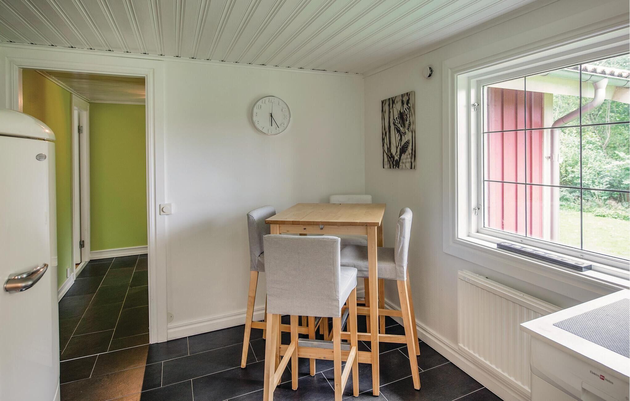 A dining area with a wooden table and four chairs, a clock on the wall, and a window with a view of greenery.