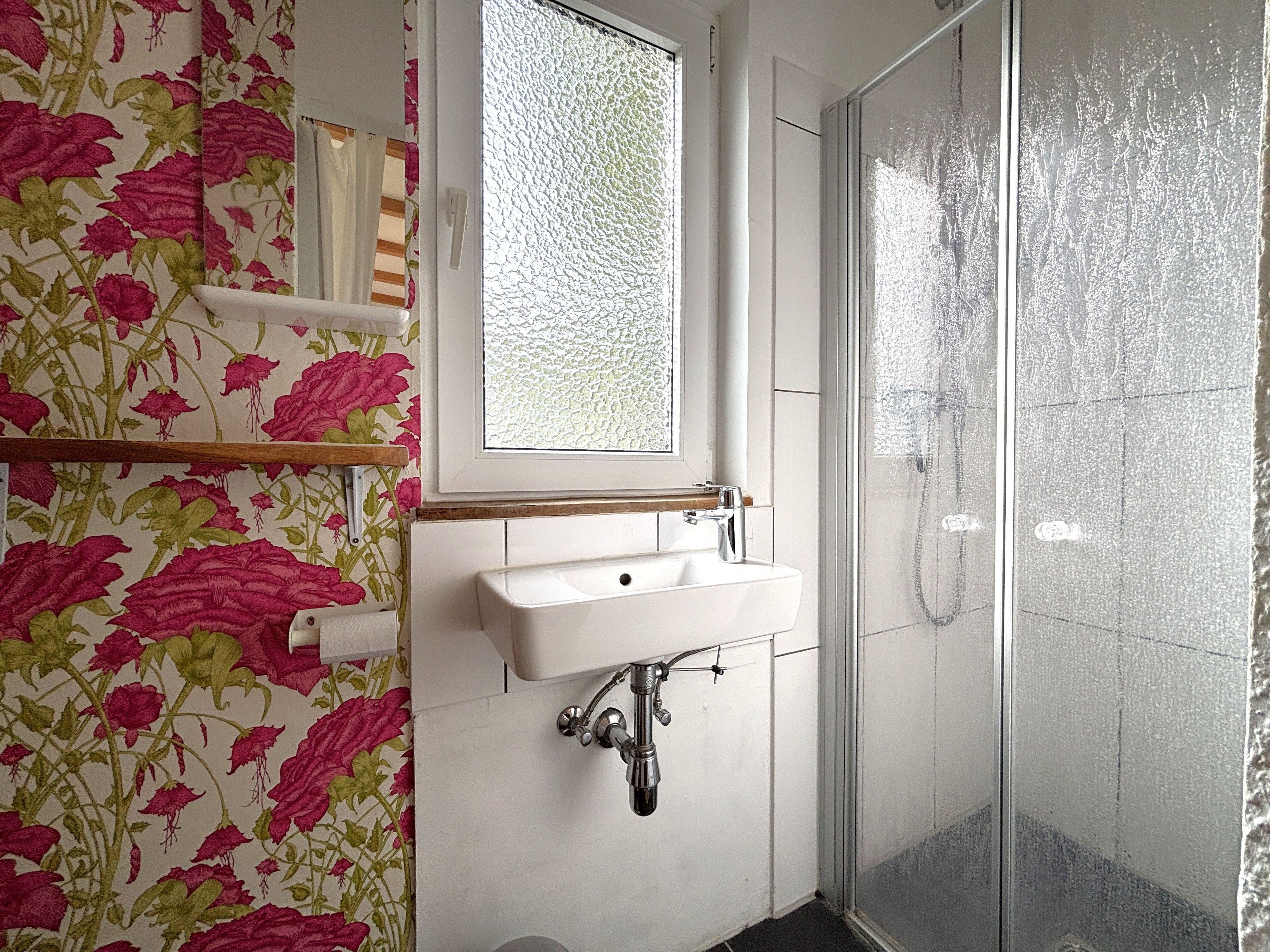 Ein Badezimmer mit floralem Wandpapier, einem weißen Waschbecken und einer Duschkabine aus Glas.