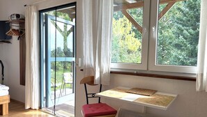 A room with a wooden floor, a desk with a chair, a window with curtains, and a view of trees outside.