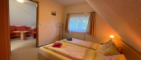Un dormitorio con dos camas, una ventana con cortinas, un radiador, una mesita y un sofá rojo en la habitación contigua.
