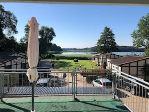 A view from a balcony overlooking a lake, with a building labeled 'Ferlenhäuser' and a parking area with cars.