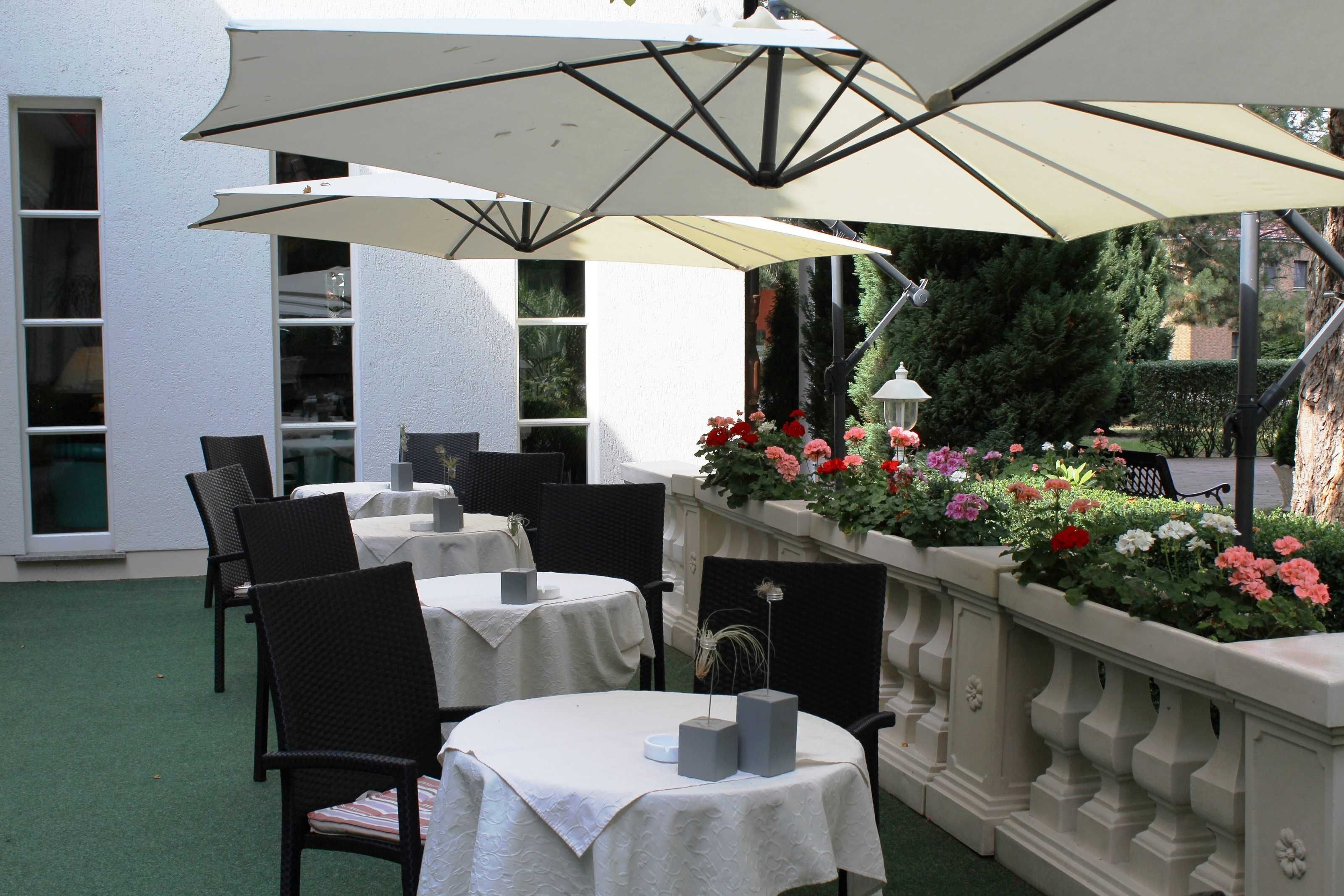 Eine Terrassenanlage im Freien mit Tischen und Stühlen, beschattet durch große Sonnenschirme, und ein Blumenbeet mit verschiedenen Blumen.