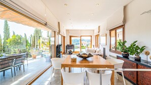 A spacious dining area with a wooden table and white chairs, large windows offering a view of greenery and buildings, and a balcony with outdoor seating. - Stunning home in Mancor de la Vall (Mancor de la Vall)