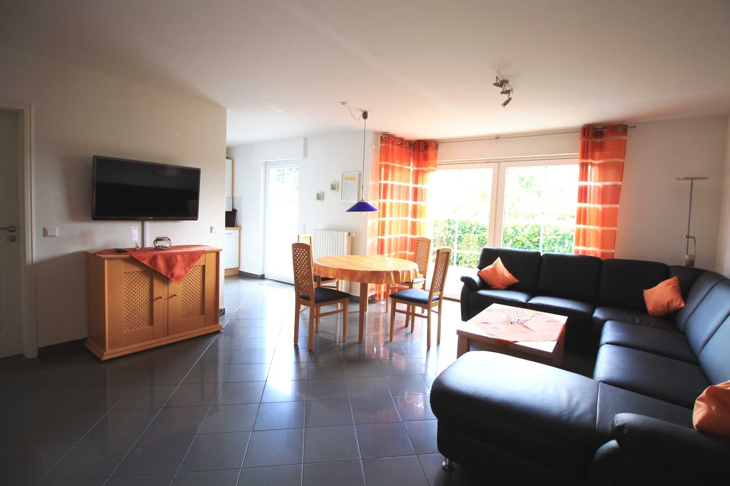 A living room with a black sofa, a wooden dining table with chairs, a television on the wall, and a window with curtains.