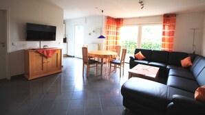 Un salon avec un canapé noir, une table à manger en bois avec des chaises, un téléviseur fixé au mur et une fenêtre avec des rideaux.