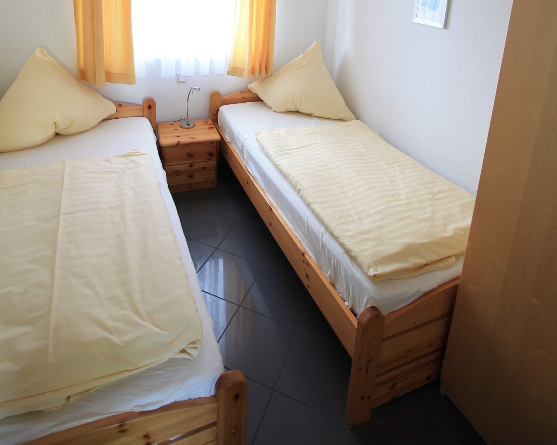Een kleine, schone kamer met twee houten bedden, een nachtkastje en een raam met gordijnen.