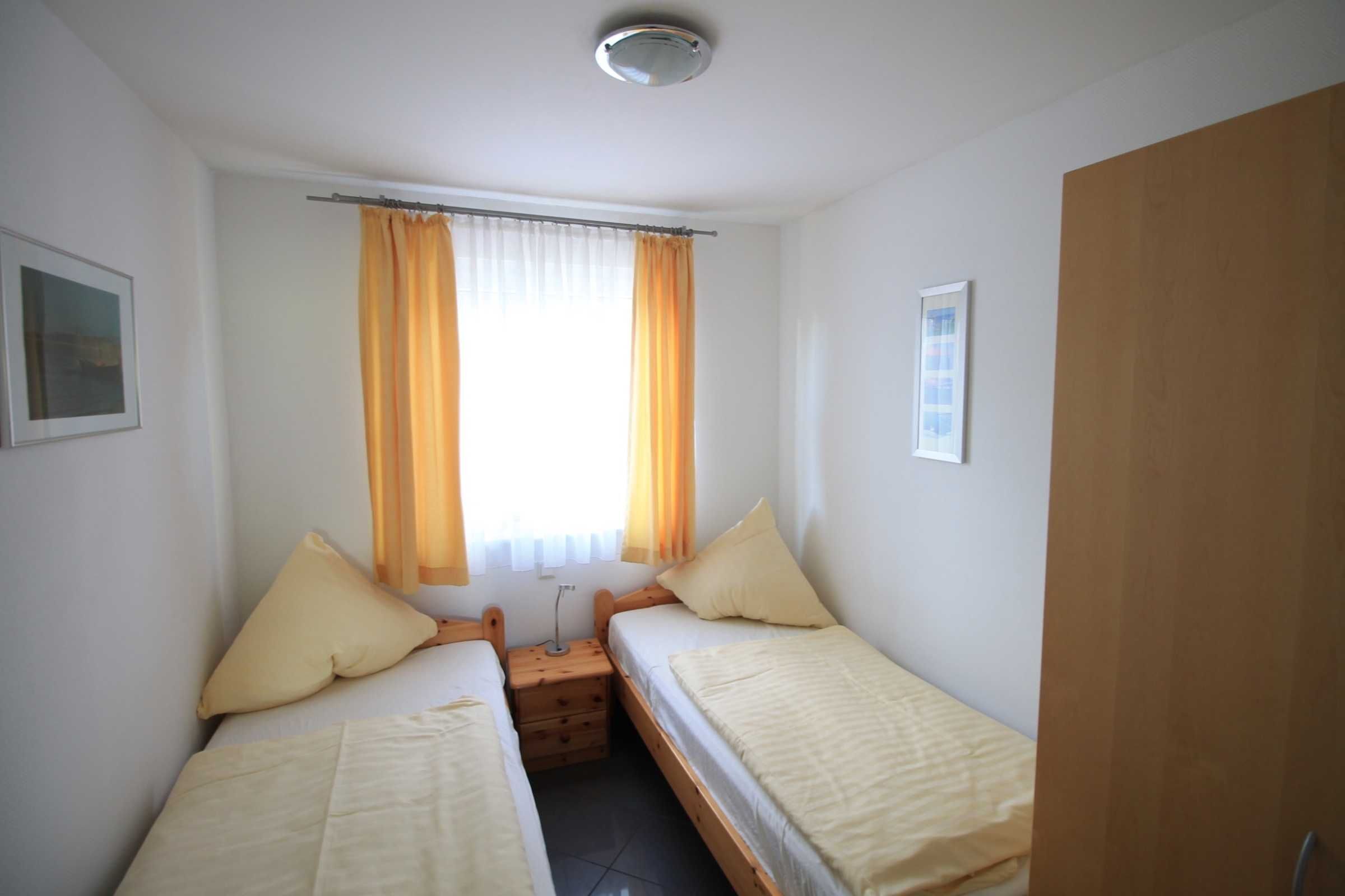 Una habitación pequeña y limpia con dos camas individuales, una mesita de noche, una ventana con cortinas y una puerta.
